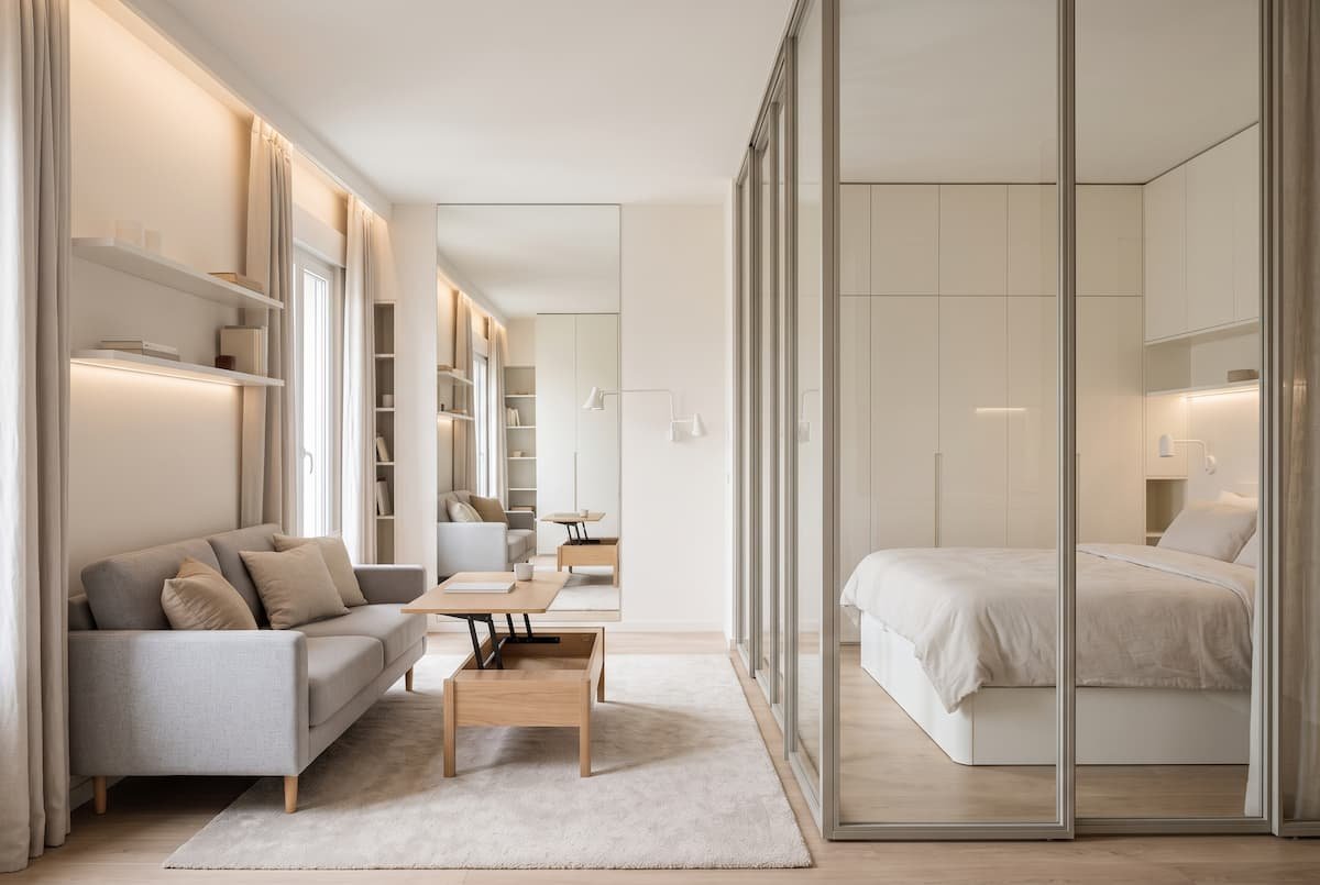 `Un pequeño apartamento tipo estudio con un cerramiento de vidrio con perfilería fina de metal que divide la zona de estar de la zona de dormitorio sin cortar la luz natural ni la visibilidad. A la izquierda se observa un sofá gris, una mesa de centro elevable de madera sobre una alfombra clara y estanterías integradas, mientras que a la derecha se aprecia una cama de tonos claros y armarios empotrados, todo en un ambiente cálido y ordenado.